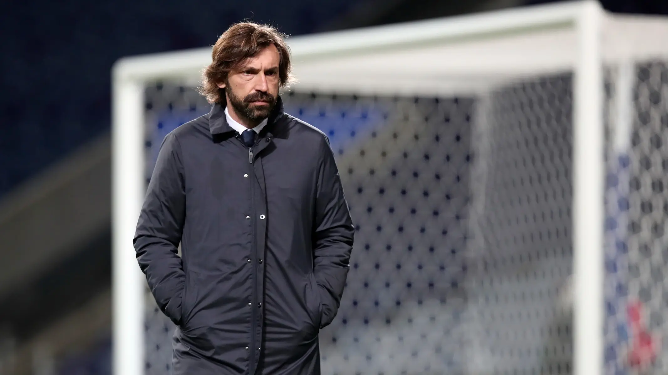 Andrea Pirlo standing on a football pitch in front of a goal net wearing a dark blue jacket