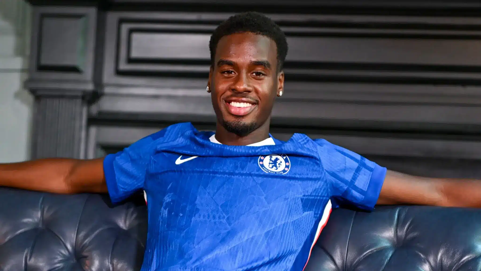 Jamie Gittens smiling while wearing a blue Chelsea FC jersey, sitting on a leather sofa.